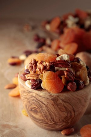Foto de Dried fruits and nuts on a beige ceramic table. The mix of nuts, apricots, and raisins in a wooden bowl. - Imagen libre de derechos