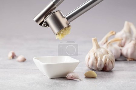 Foto de Garlic on a grey stone table. Squeeze the garlic with a garlic press into a small bowl. - Imagen libre de derechos
