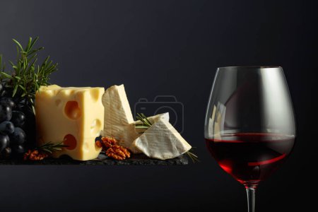 Foto de Vino tinto con queso, nueces, uvas y romero sobre fondo negro. - Imagen libre de derechos