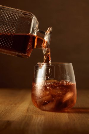 Téléchargez les photos : Le whisky est versé dans un verre barré avec de la glace naturelle. Verre de whisky sur une table en chêne. - en image libre de droit