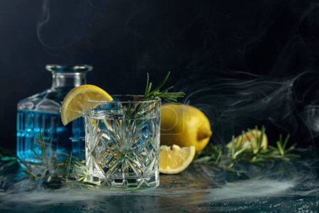 Foto de Cóctel Gin-tonic con limón y romero en una copa de cristal. El romero ahumado el coctel a la antigua sobre la mesa. - Imagen libre de derechos