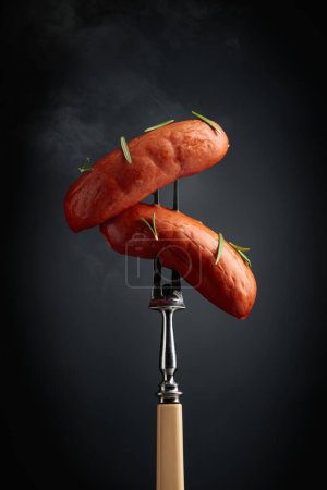 Foto de Salchichas hervidas con romero en un tenedor. Salchichas calientes con humo sobre fondo negro. - Imagen libre de derechos