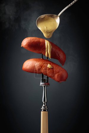 Foto de Salchichas hervidas con mostaza en un tenedor. Salchichas calientes con humo sobre fondo negro. - Imagen libre de derechos
