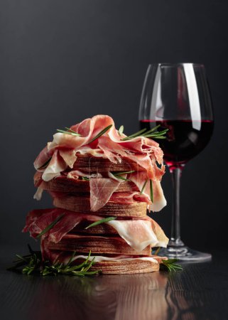 Photo for Spanish jamon or Italian prosciutto with bread, rosemary, and red wine on a black wooden table. - Royalty Free Image