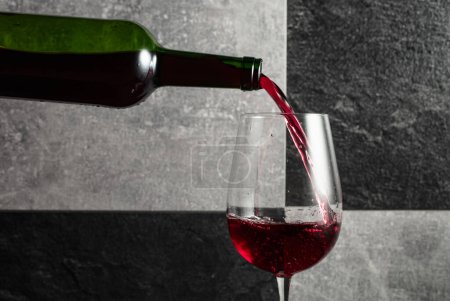Téléchargez les photos : Le vin rouge est versé de la bouteille dans un verre. Fond en pierre grise et noire. Espace de copie. - en image libre de droit