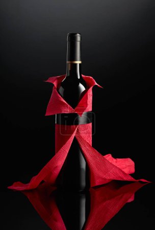 Foto de Botella de vino tinto envuelta en papel crepé sobre fondo negro. La botella parece una mujer con un vestido de noche rojo. - Imagen libre de derechos