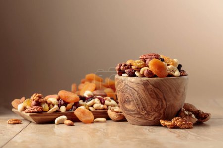 Téléchargez les photos : Dried fruits and nuts on a beige ceramic table. The mix of nuts, apricots, and raisins in a wooden bowl. Copy space. - en image libre de droit
