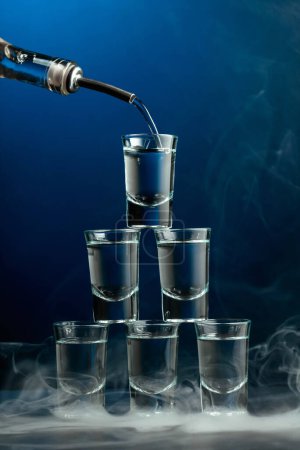 Téléchargez les photos : Lunettes de vodka en fumée sur fond bleu foncé. Concept de boissons club. - en image libre de droit