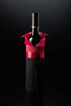 Photo for Bottle of red wine wrapped in crepe paper on a black background. The bottle looks like a man in a red shirt. - Royalty Free Image