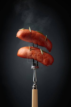 Téléchargez les photos : Saucisses bouillies avec romarin sur une fourchette. Saucisses chaudes avec de la fumée sur fond noir. - en image libre de droit