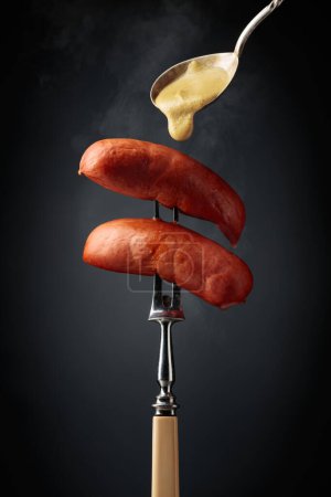 Foto de Boiled sausages with mustard on a fork. Hot sausages with smoke on a black background. - Imagen libre de derechos