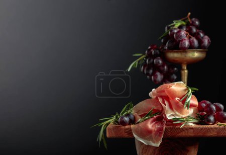 Foto de Prosciutto con uvas y romero sobre una mesa de madera. Copiar espacio. - Imagen libre de derechos