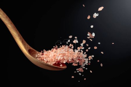 Téléchargez les photos : Le sel rose de l'Himalaya est versé avec une cuillère en bois. Himalaya sel sur un fond sombre. Espace de copie. - en image libre de droit