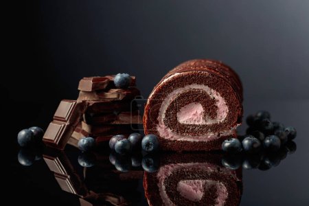 Foto de Chocolate roll cake with blueberries and a broken black chocolate bar on a black reflective background. - Imagen libre de derechos