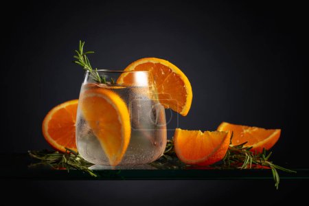 Foto de Cóctel con hielo, naranja y romero en un vaso congelado. Una bebida refrescante helada en vidrio nebulizado sobre un fondo negro. - Imagen libre de derechos