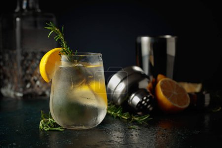 Foto de Cóctel gin tonic con hielo, limón y romero. Una bebida refrescante helada en vidrio nebulizado sobre un fondo oscuro. - Imagen libre de derechos