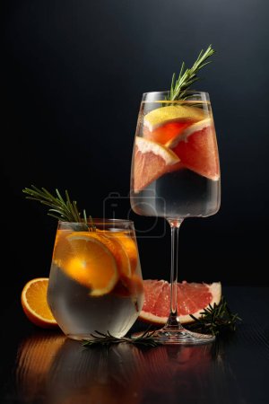 Foto de Gin tonic con hielo, romero, naranja y pomelo en vasos esmerilados. Gafas con cócteles en una mesa de madera negra. - Imagen libre de derechos