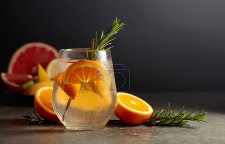 Foto de Cóctel gin tonic con hielo, romero y naranja sobre una mesa de piedra. En el fondo hay varios cítricos para hacer cócteles. - Imagen libre de derechos