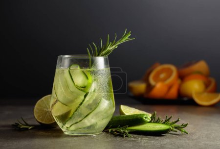 Photo for Cocktail gin tonic with ice, rosemary, lime, and cucumber on a stone table. In the background are various citrus fruits for making cocktails. - Royalty Free Image