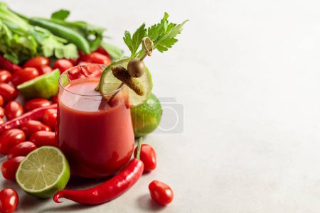 Foto de Cóctel Bloody Mary decorado con pepinillo, olivo, lima y apio. Bebida de tomate con ingredientes en una mesa de piedra blanca. - Imagen libre de derechos