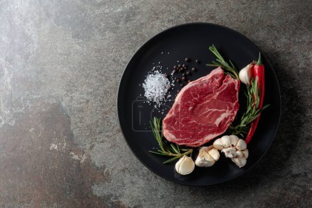 Photo for Raw beef steak for grilling with rosemary, garlic, salt, and pepper. Old stone background. Top view. - Royalty Free Image