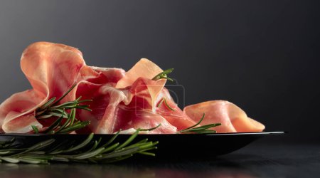 Photo for Italian prosciutto or Spanish jamon with rosemary on a black plate. - Royalty Free Image