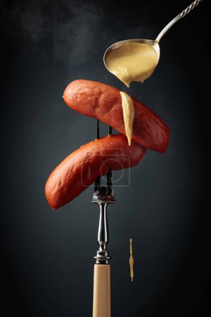 Foto de Salchichas hervidas con mostaza en un tenedor. Salchichas calientes con humo sobre fondo negro. - Imagen libre de derechos
