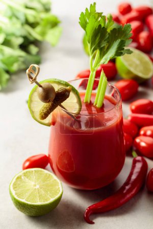Foto de Cóctel Bloody Mary decorado con pepinillo, olivo, lima y apio. Bebida de tomate con ingredientes en una mesa de piedra blanca. - Imagen libre de derechos