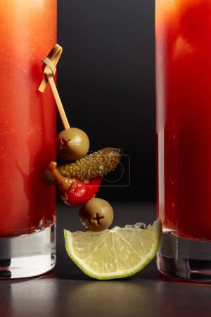 Foto de Cóctel Bloody Mary con pepinillo, aceituna, lima y pimiento rojo. Bebida de tomate sobre fondo negro. - Imagen libre de derechos