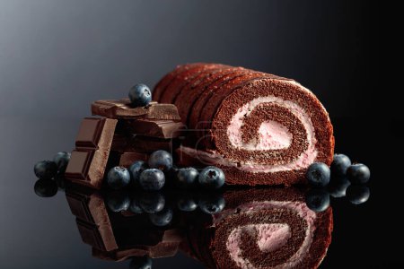 Téléchargez les photos : Chocolate roll cake with blueberries and a broken black chocolate bar on a black reflective background. - en image libre de droit