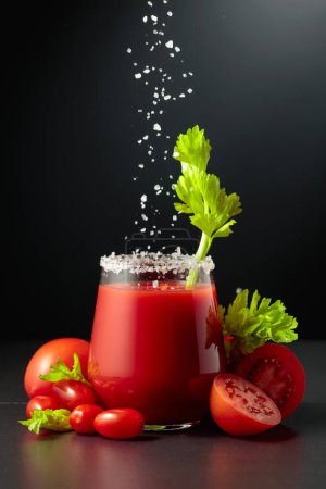 Foto de La sal marina se vierte en un vaso de jugo de tomate con apio. El vaso de jugo y verduras frescas están sobre un fondo negro. - Imagen libre de derechos