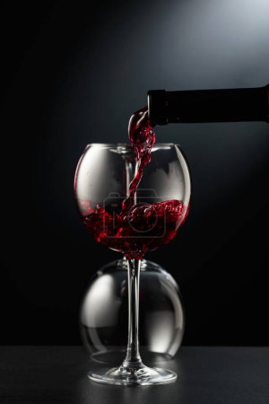 Téléchargez les photos : Verser le vin rouge dans un verre de vin sur un fond noir. Un verre renversé est en arrière-plan. - en image libre de droit