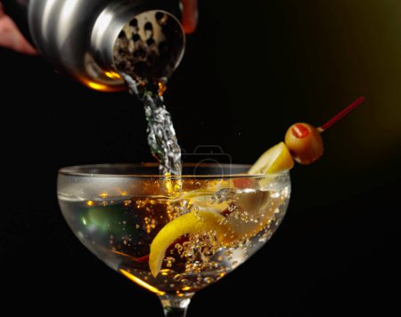 Martini mit grünen Oliven und Zitrone. Der Cocktail wird aus einem Shaker in ein Glas gegossen.
