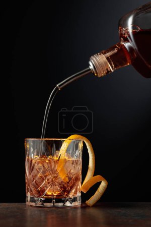 Foto de Cóctel a la antigua con hielo y piel de naranja. Whisky se vierte en un vaso con hielo. - Imagen libre de derechos