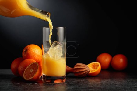 Téléchargez les photos : Verser le jus d'orange d'une bouteille dans un verre avec de la glace. Fond noir avec espace de copie. - en image libre de droit