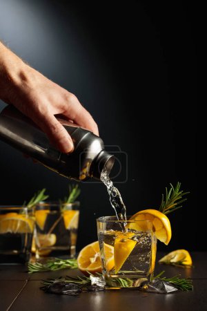 Téléchargez les photos : Cocktail Gin and Tonic au citron et romarin. Le barman verse un cocktail d'un shaker dans un verre. Espace de copie. - en image libre de droit