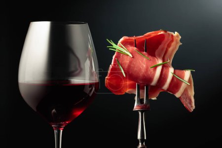 Téléchargez les photos : Verre de vin rouge et de prosciutto tranché avec romarin sur une fourchette, fond noir. - en image libre de droit
