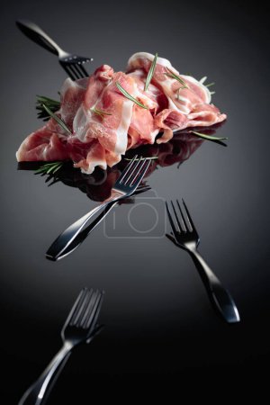 Téléchargez les photos : Prosciutto italien ou jamon espagnol au romarin. Viande et fourchettes sur fond réfléchissant noir. - en image libre de droit