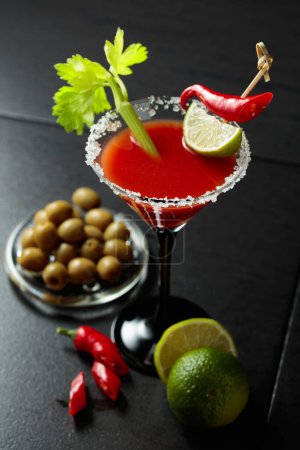 Foto de Cóctel Bloody Mary con apio, lima, pimiento rojo y aceitunas verdes. El vidrio está decorado con sal marina. - Imagen libre de derechos