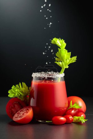 Foto de La sal marina se vierte en un vaso de jugo de tomate con apio. El vaso de jugo sobre un fondo negro. - Imagen libre de derechos