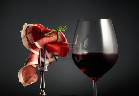 Photo for Glass of red wine and sliced prosciutto with rosemary on a fork, black background. - Royalty Free Image