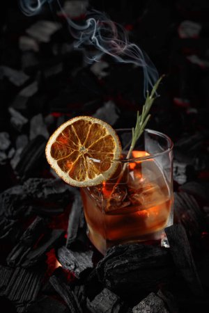 Foto de Cóctel a la antigua con hielo, rebanada de naranja seca y romero. Whisky con romero y hermosos remolinos de humo sobre un fondo de carbón quemado. - Imagen libre de derechos