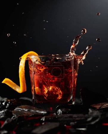 Photo for Piece of ice falls into the glass with a cocktail. Cocktail with ice and orange peel on burning charcoal. - Royalty Free Image