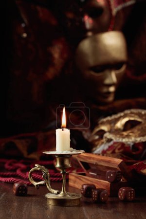Photo for Burning candle, dice, and carnival masks on an old wooden table. - Royalty Free Image