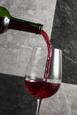 Téléchargez les photos : Le vin rouge est versé de la bouteille dans un verre. Fond en pierre grise et noire. - en image libre de droit