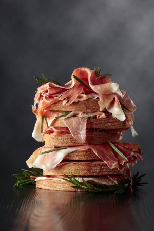 Photo for Italian prosciutto or Spanish jamon with bread and rosemary on a black wooden table. Copy space. - Royalty Free Image