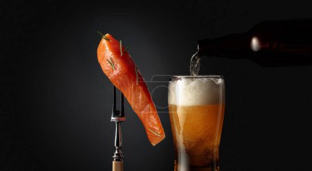 Photo for Smocked salmon with rosemary on a fork and a glass of beer on a dark background. Beer is poured from a bottle into a glass. Copy space. - Royalty Free Image