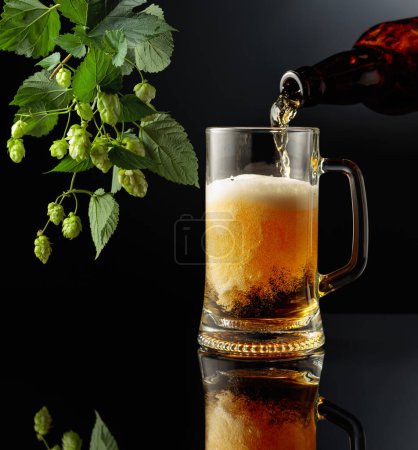 Foto de Verter cerveza de una botella en una taza. Taza de cerveza y rama de lúpulo sobre un fondo negro reflectante. Copiar espacio. - Imagen libre de derechos