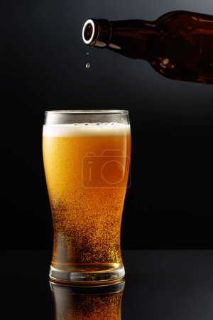 Téléchargez les photos : Verser la bière d'une bouteille dans un verre. Verre de bière sur fond réfléchissant noir. - en image libre de droit