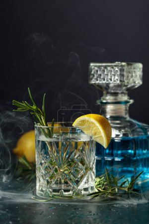 Foto de Cóctel Gin-tonic con limón y romero en una copa de cristal. El romero ahumado el coctel a la antigua sobre la mesa. - Imagen libre de derechos